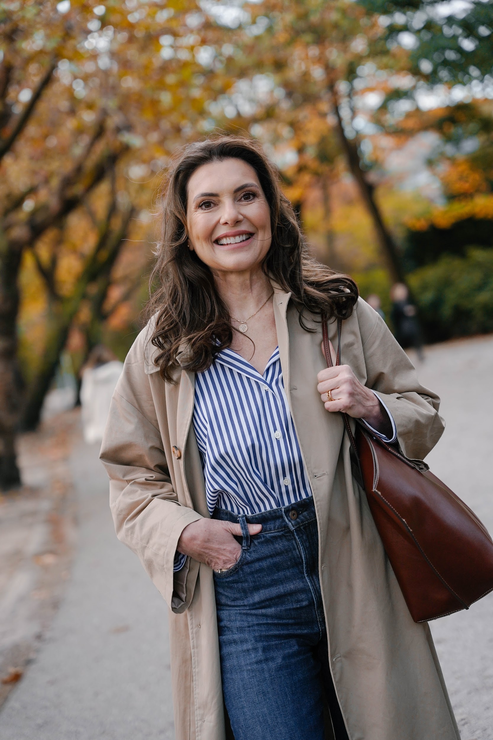 Marjena in een blauwe blouse, kijkt lachend in de camera. Bomen in herfstkleuren op de achtergrond